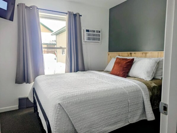 A bedroom with a bed, curtains, a wall-mounted air conditioner, and a view of the outside. - Scotty's Motel (Munising)