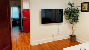Una habitación con una televisión de pantalla plana montada en la pared, suelo de madera, un refrigerador rojo y una planta en maceta.