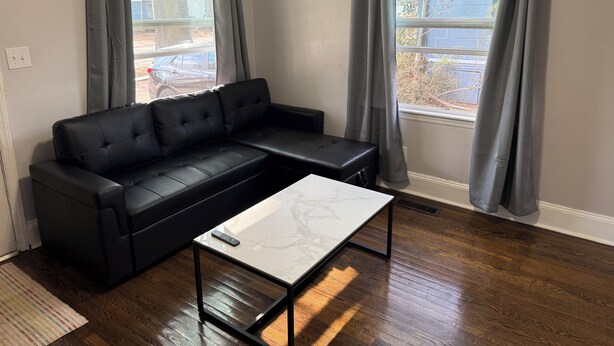 Una sala de estar moderna con un sofá seccional de cuero negro, una mesa de centro de vidrio y dos ventanas con cortinas grises.
