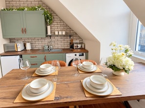 A dining area with a wooden table set for four, a kitchen in the background with a brick wall, and a window with a view. - Central Ayr Attic Apartment + Panoramic Town Views (Ayr)