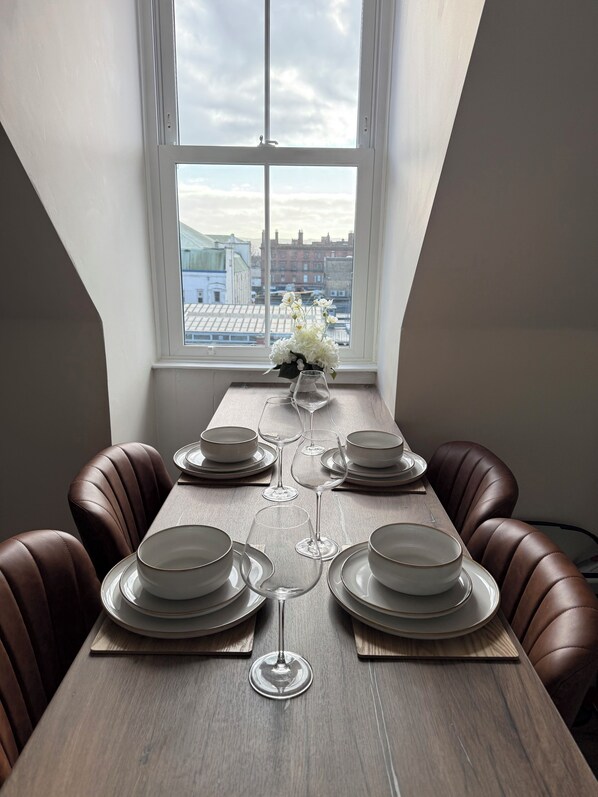 A dining table set with plates, bowls, and wine glasses, positioned in front of a window with a cityscape view.