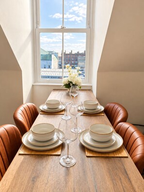 A dining table set with plates, bowls, and wine glasses, positioned in front of a window with a cityscape view. - Central Ayr Attic Apartment + Panoramic Town Views (Ayr)