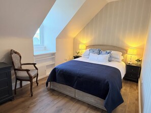 A bedroom with a sloped ceiling, a bed with a dark blue bedspread, two bedside lamps, a wooden chair, and a nightstand with a lamp.