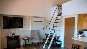 Espaço compacto com televisão, uma pequena mesa com uma planta, uma cadeira, uma escada e uma cozinha com banquetas.