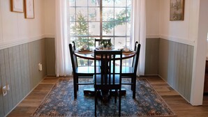 Uma sala de jantar com uma mesa posta para quatro pessoas, um tapete estampado e uma janela grande com cortinas.
