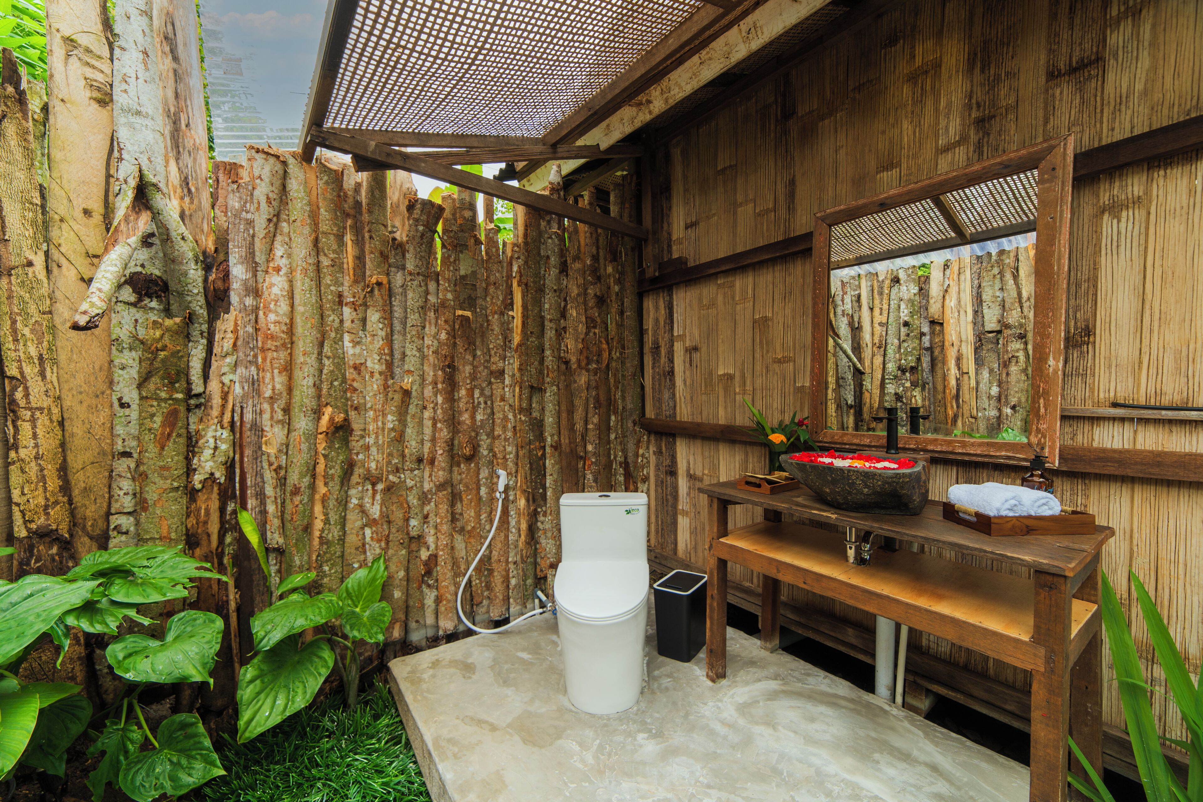 A rustic bathroom with a wooden wall, a white toilet, a sink with a mirror, and a small trash bin.