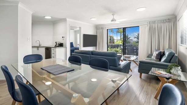 A modern living room with a glass dining table, blue chairs, a grey sofa, and a small coffee table. - Rare 4BR Ground Floor Exclusive Apt (Mooloolaba)