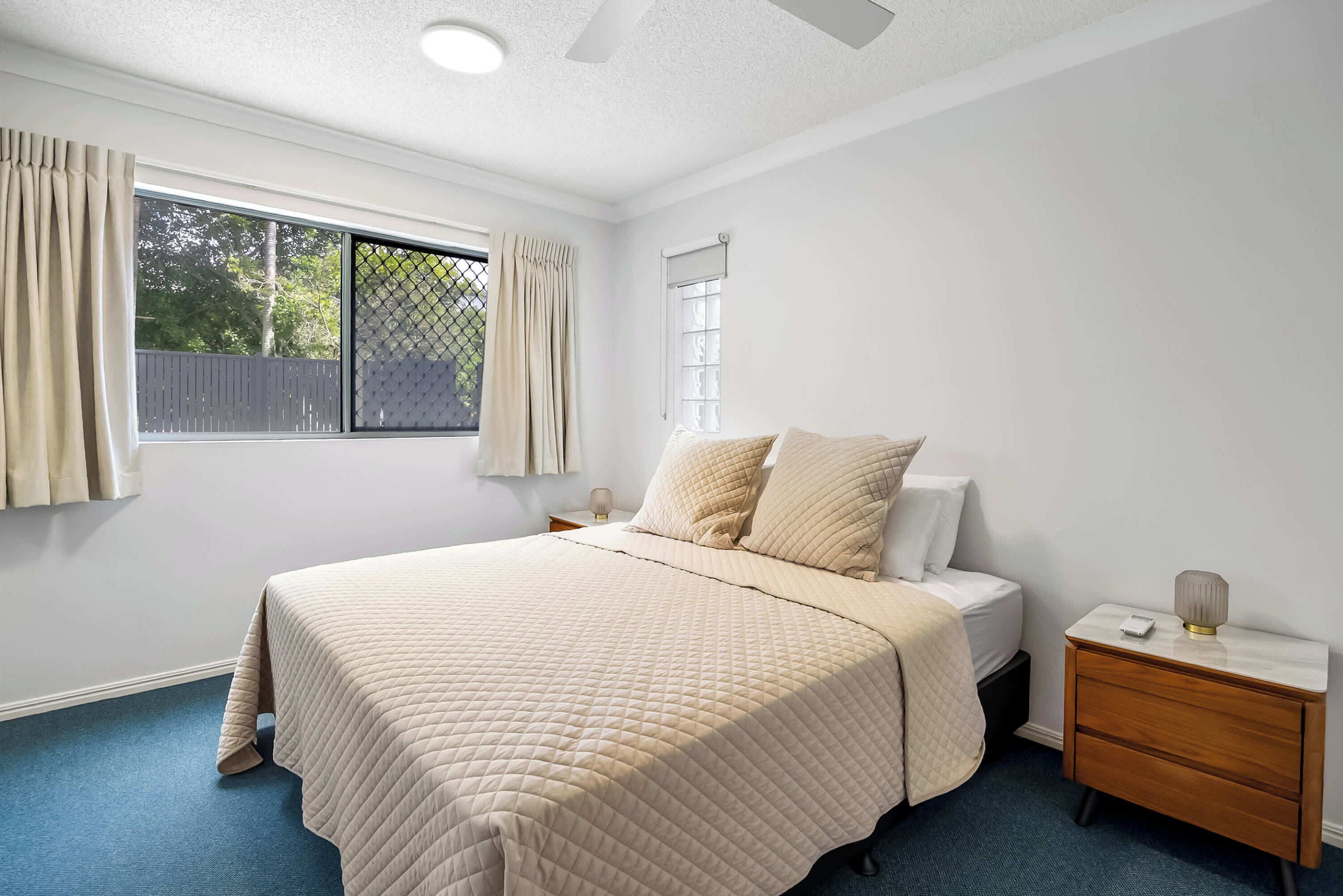 Une chambre à coucher avec un lit, une table de chevet, une fenêtre avec des rideaux et un ventilateur de plafond.