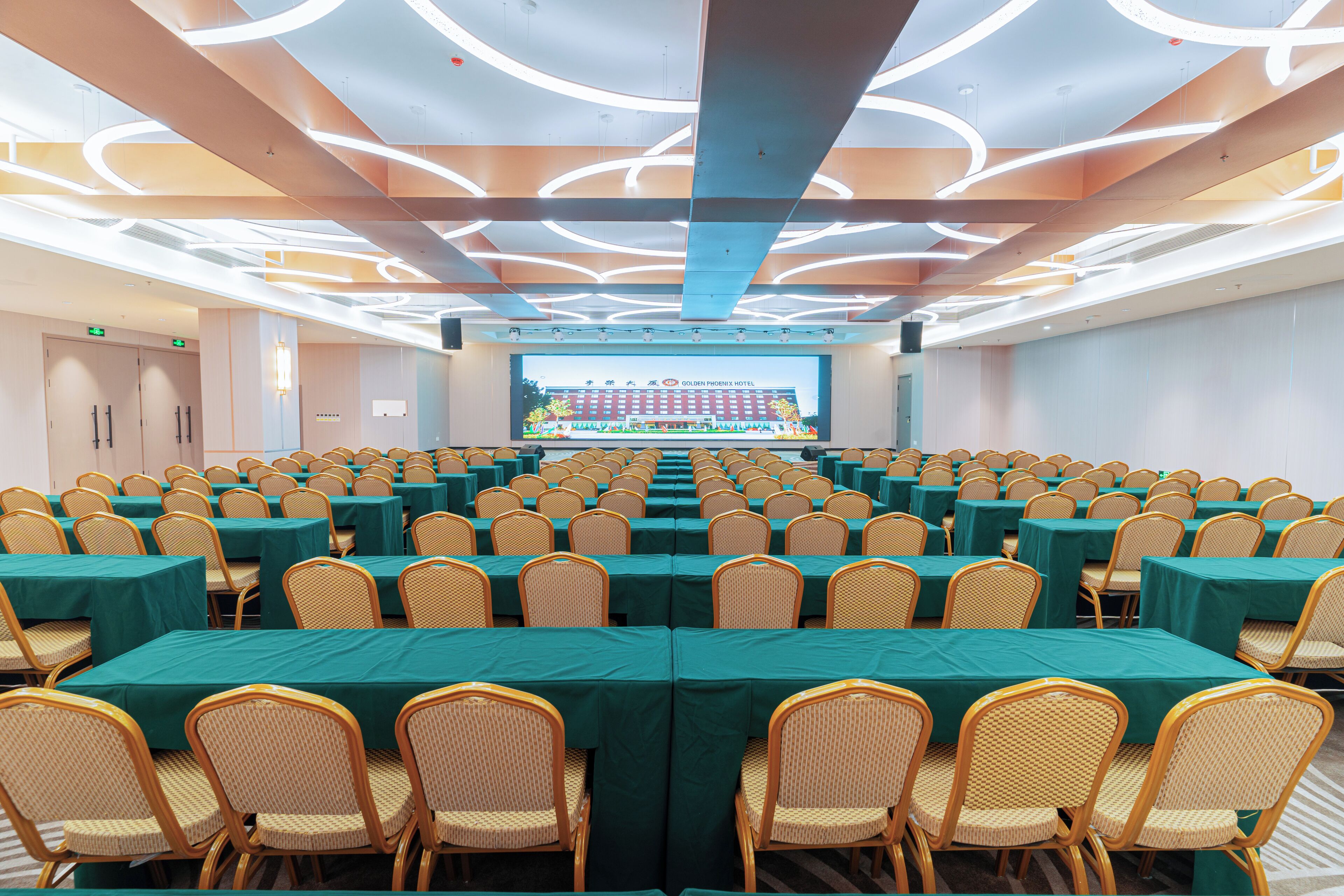 A conference room with rows of tables and chairs, a large screen at the front, and a modern ceiling design.