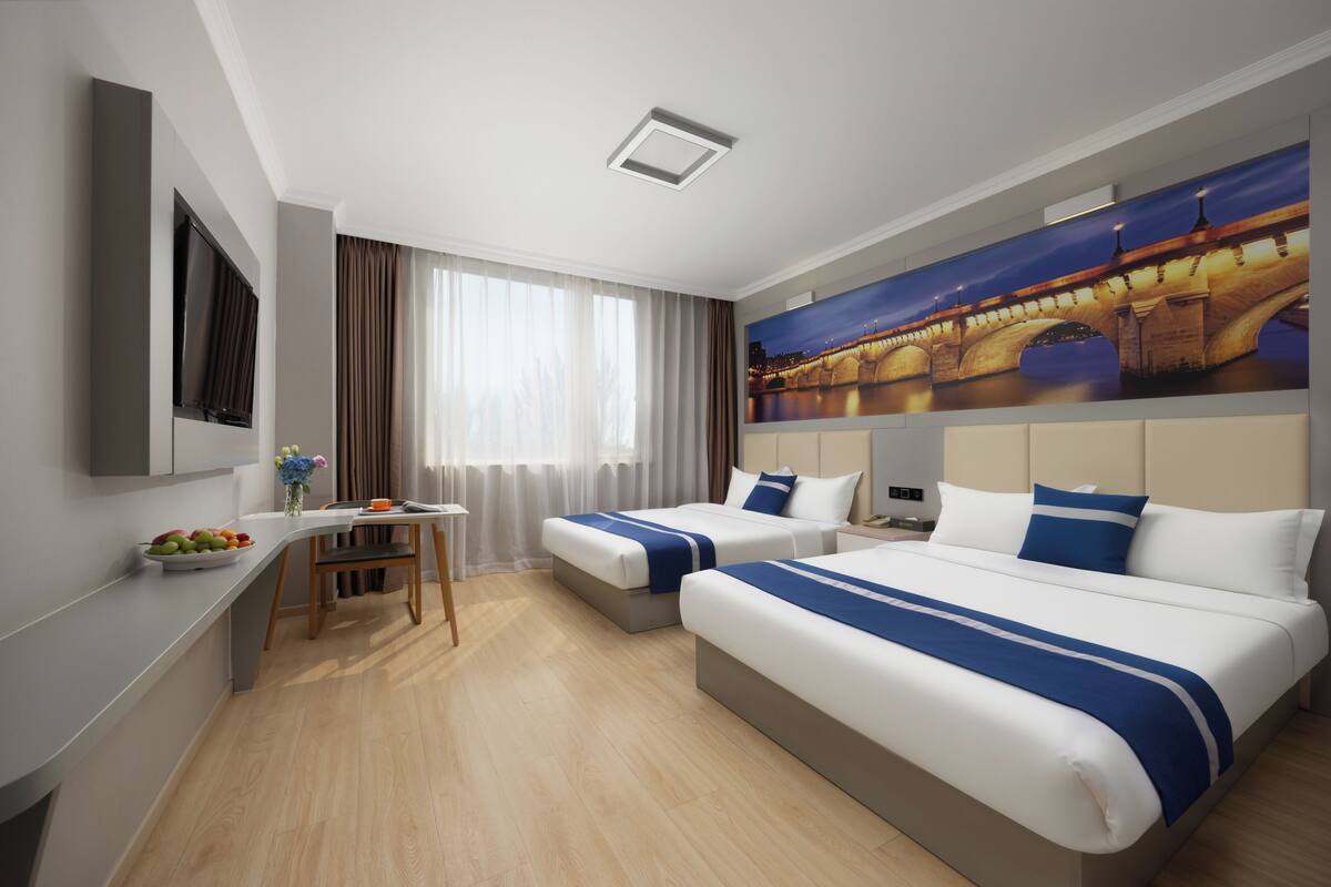 A hotel room with two beds, a desk, a chair, a fruit bowl, and a painting of a bridge.
