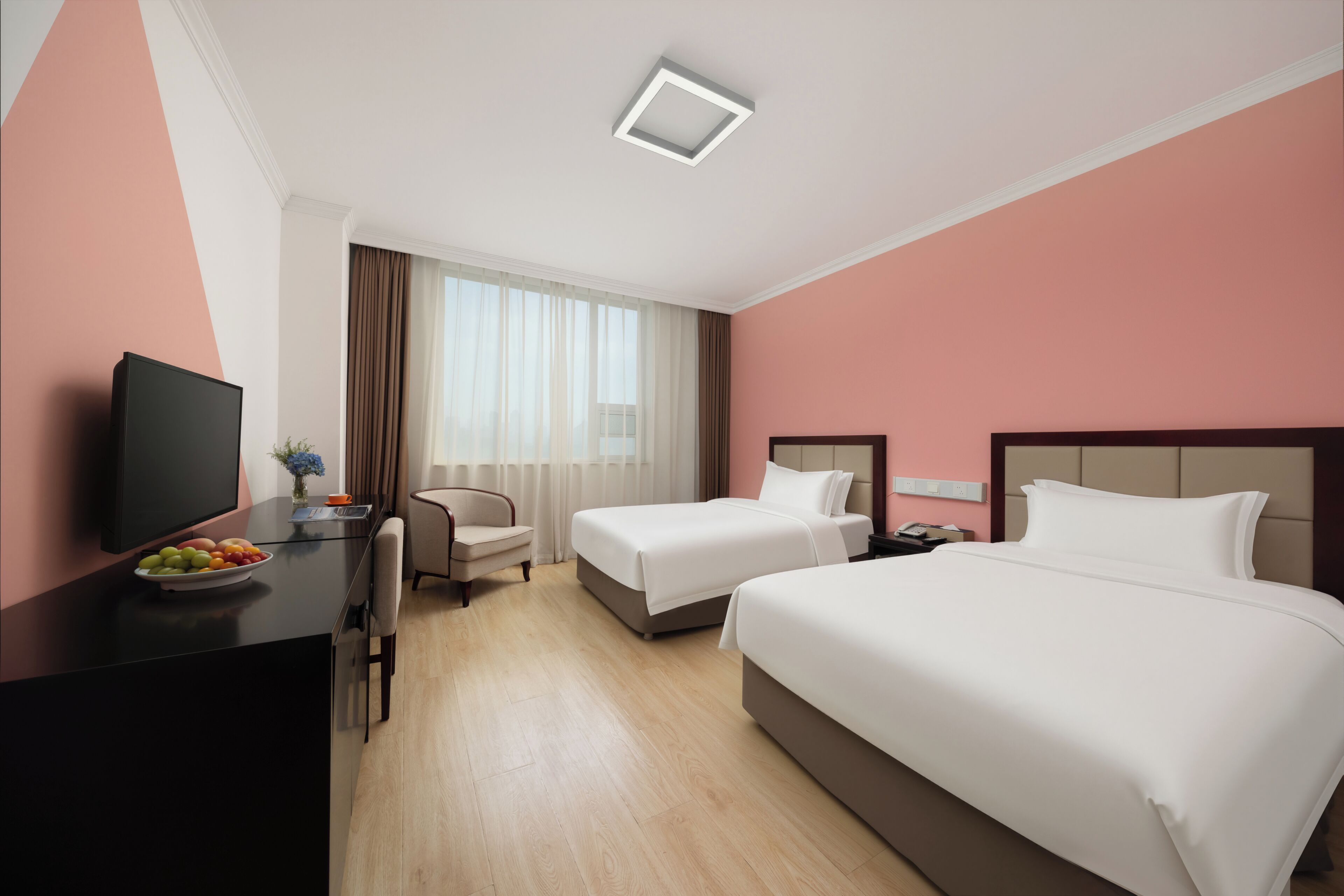 Habitación de hotel con cama, escritorio con televisor, silla, mesita con fruta y ventana con cortinas.