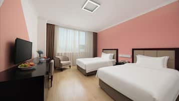 Een hotelkamer met een bed, een bureau met een televisie, een stoel, een klein tafeltje met fruit en een raam met gordijnen.