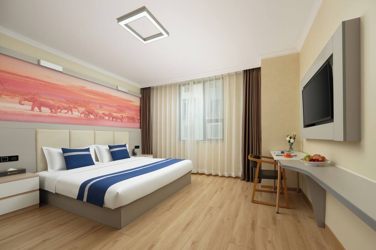 A modern hotel room with a large bed, a desk, a flat-screen TV, and a cityscape painting on the wall.