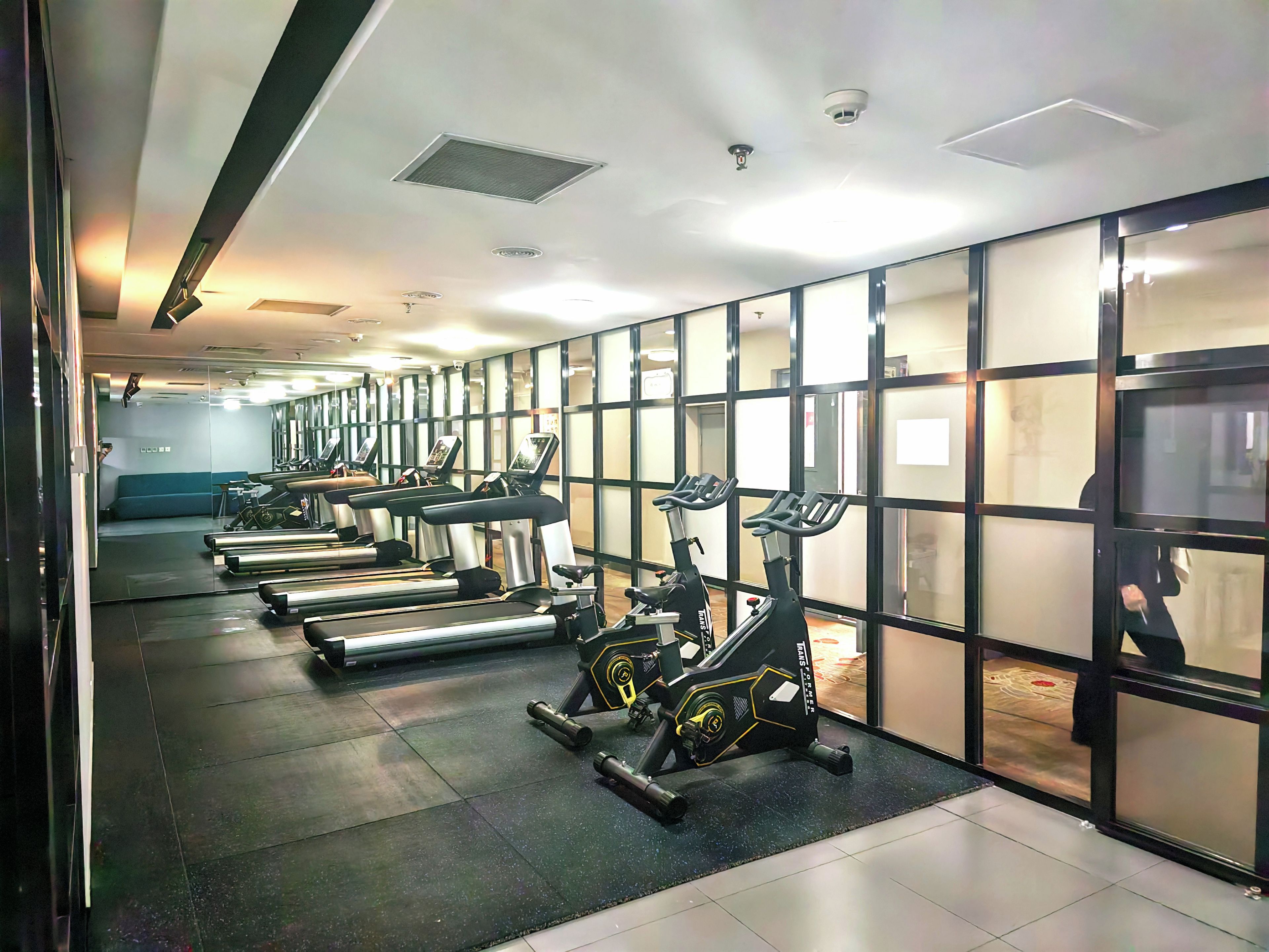 A gym with treadmills and stationary bikes arranged in rows, a mirrored wall, and a tiled floor.