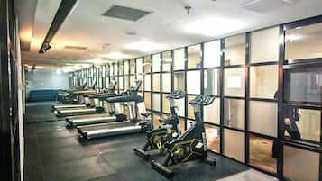 A gym with treadmills and stationary bikes arranged in rows, a mirrored wall, and a tiled floor.