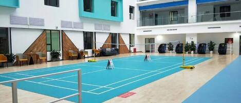 A multi-level indoor sports facility with a blue tennis court, seating areas, and a high ceiling.