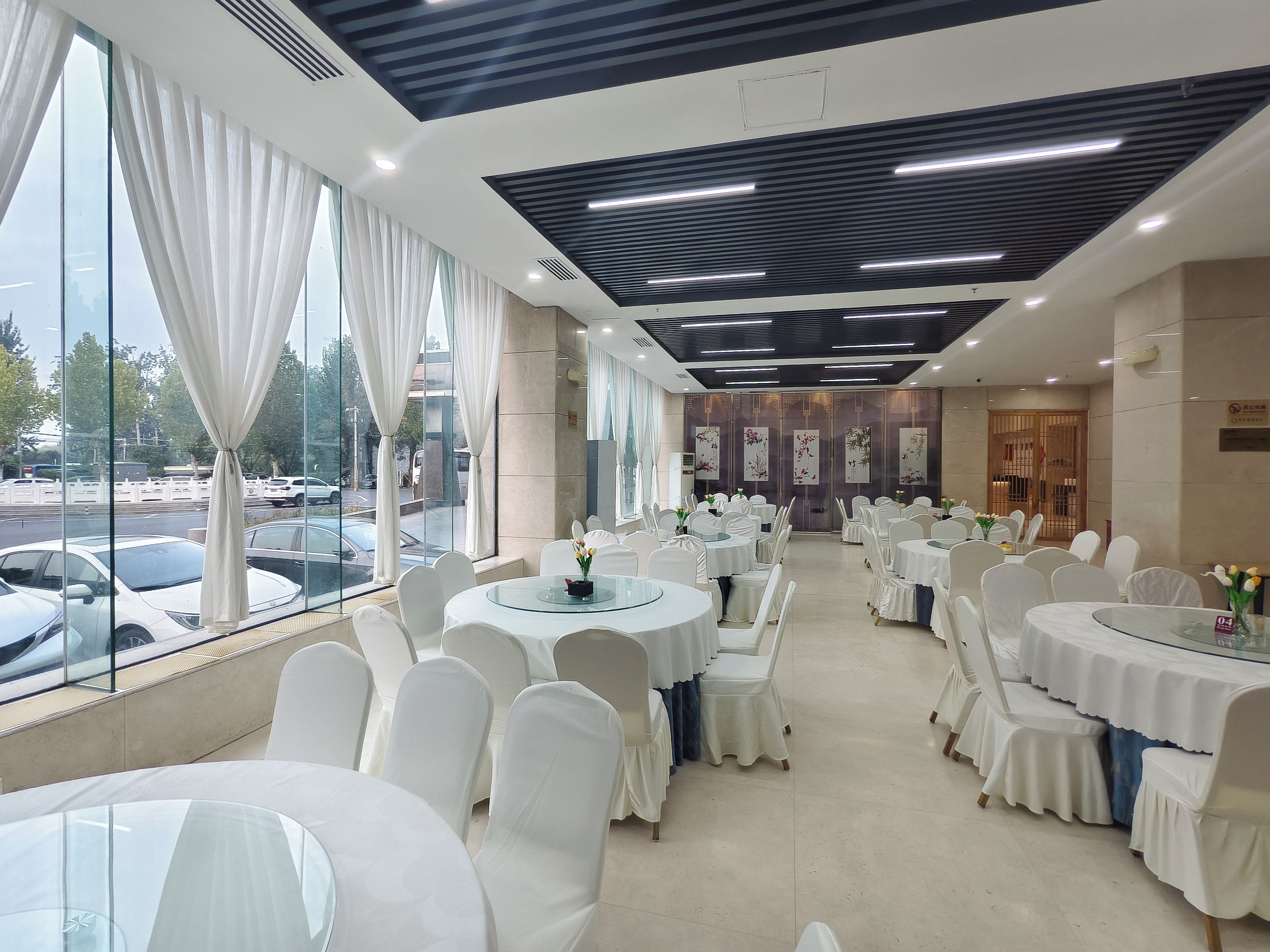 A hotel dining area with round tables, white chairs, and large windows overlooking a parking area.