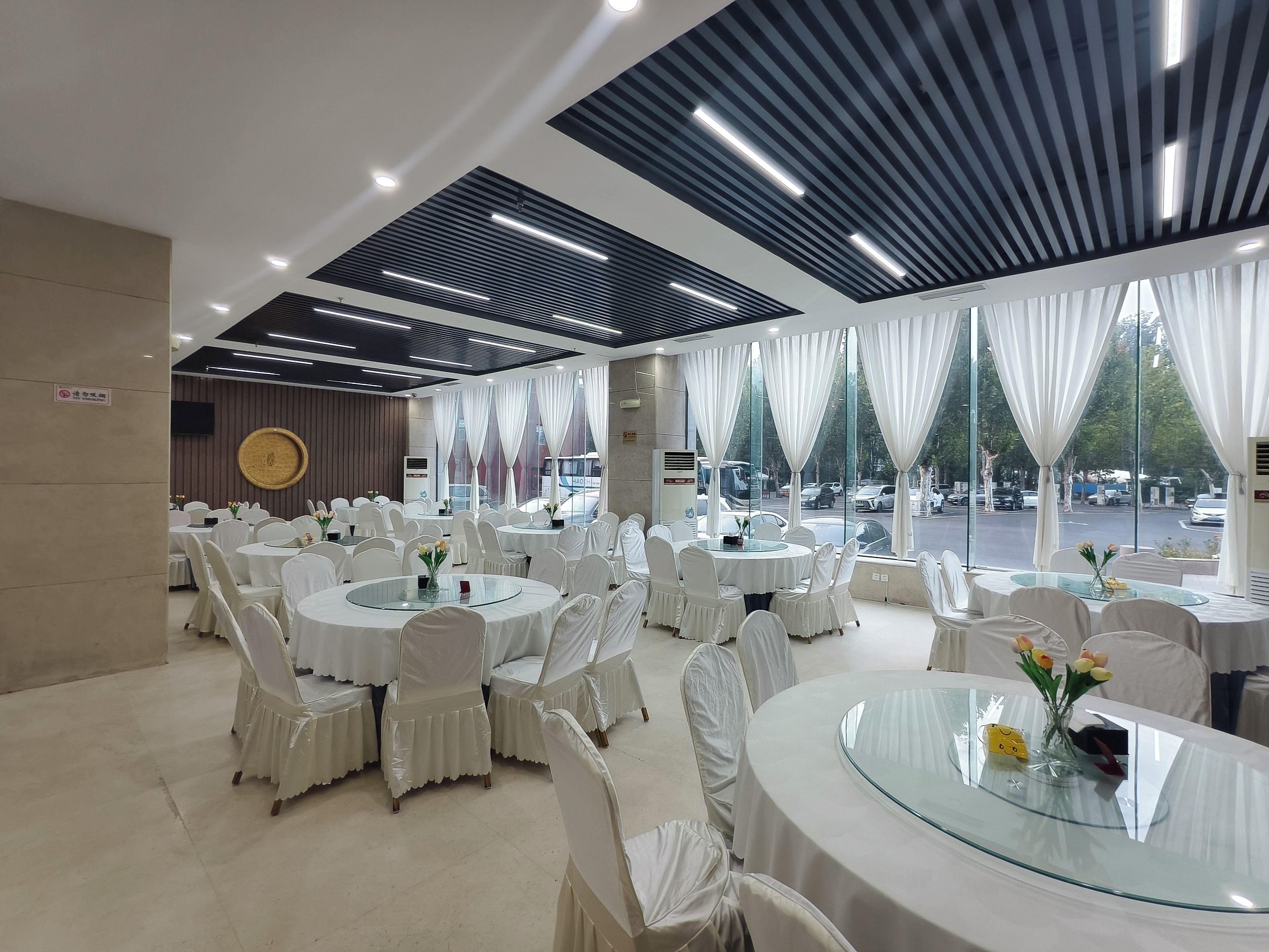 A banquet hall with round tables and chairs, large windows with curtains, and a modern ceiling design.