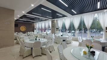 A banquet hall with round tables and chairs, large windows with curtains, and a modern ceiling design.
