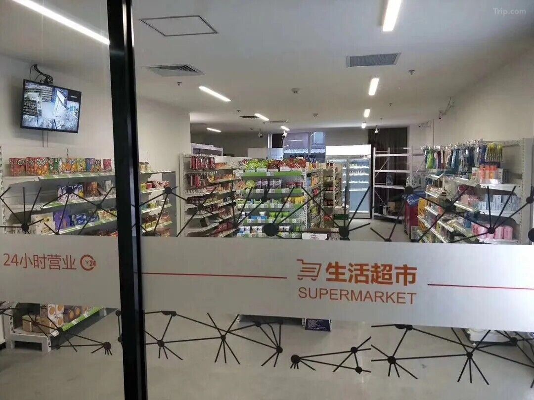 A supermarket interior with shelves stocked with various products, a 24-hour operation sign, and a security camera on the ceiling.