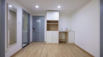 A small, well-lit room with a kitchenette, a door leading to another room, and a light-colored wooden floor.