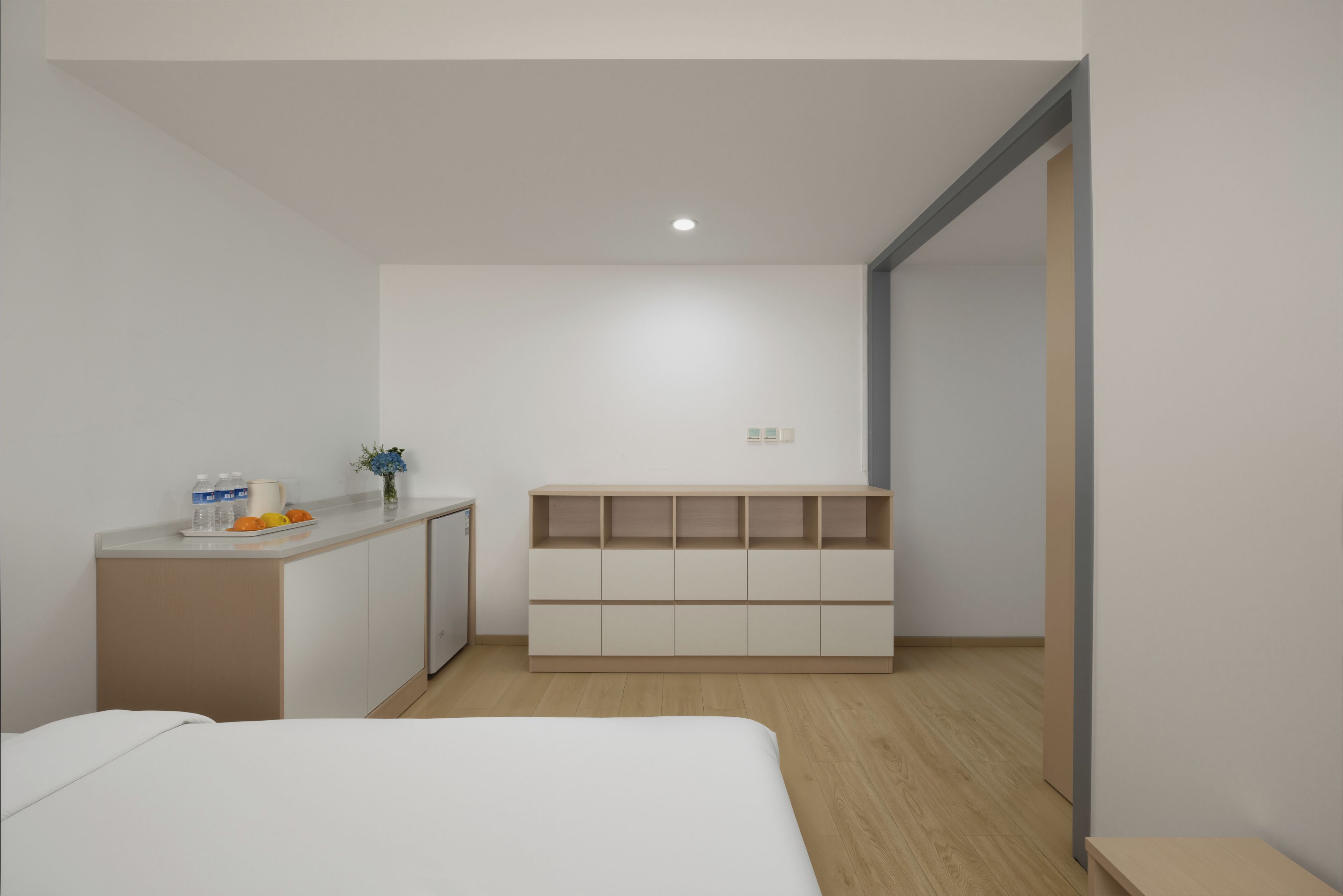 Habitación de hotel minimalista con una cama, un escritorio pequeño, un armario y estanterías integradas.