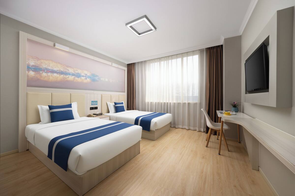 A hotel room with two beds, a desk, a TV, and a large window with curtains.