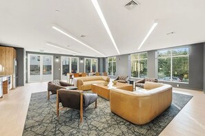 A spacious living area with a large sectional sofa, a coffee table, and a fireplace. There are multiple seating options and a view of the outdoors through large windows. - Blueground | The Village, fitness center, nr Liberty State Park (Jersey City)