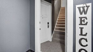 Um corredor com uma porta branca e uma escada que leva a um segundo andar. Uma placa com a palavra 'WELCOME' é visível na parede.