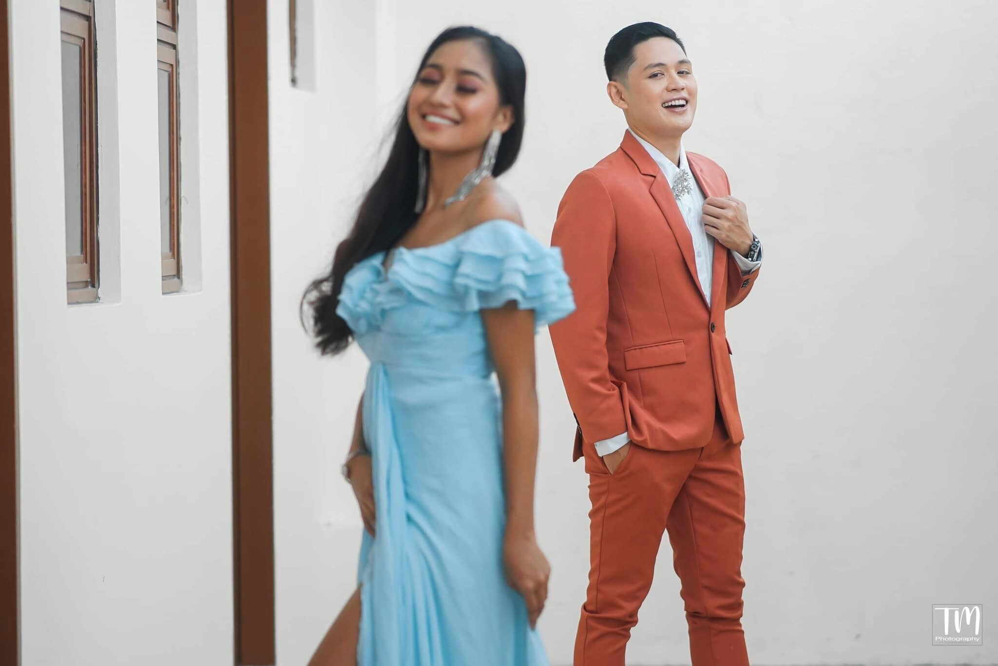 A person in a light blue dress and another in a red suit walking in a hallway with white walls and wooden doors.