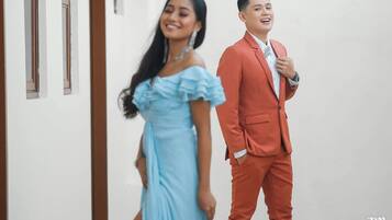 A person in a light blue dress and another in a red suit walking in a hallway with white walls and wooden doors.