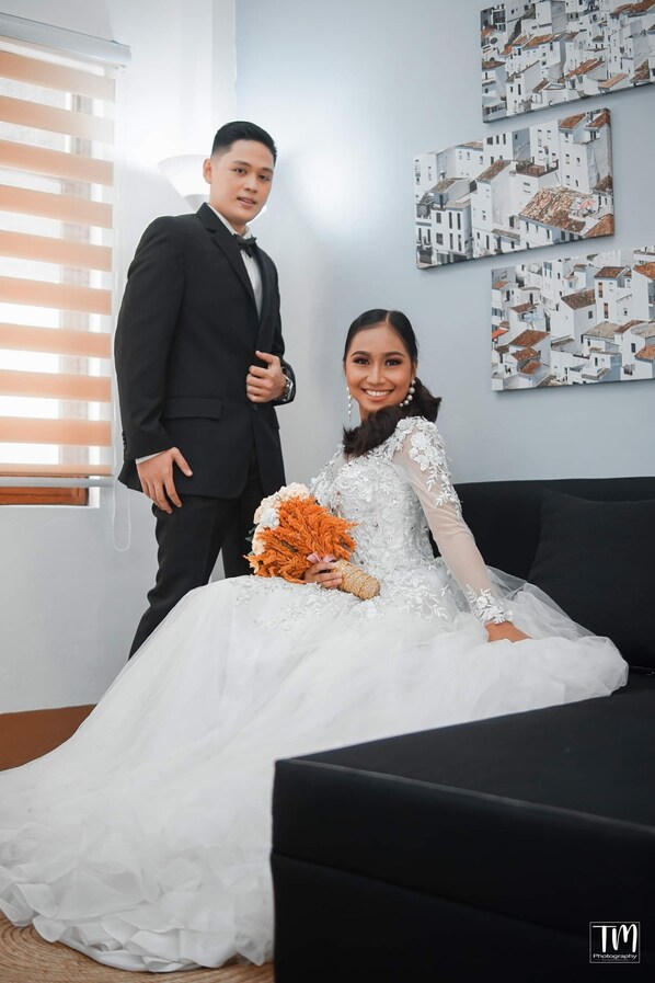 A wedding couple seated on a black couch in a room with a large window and wall art depicting white buildings.