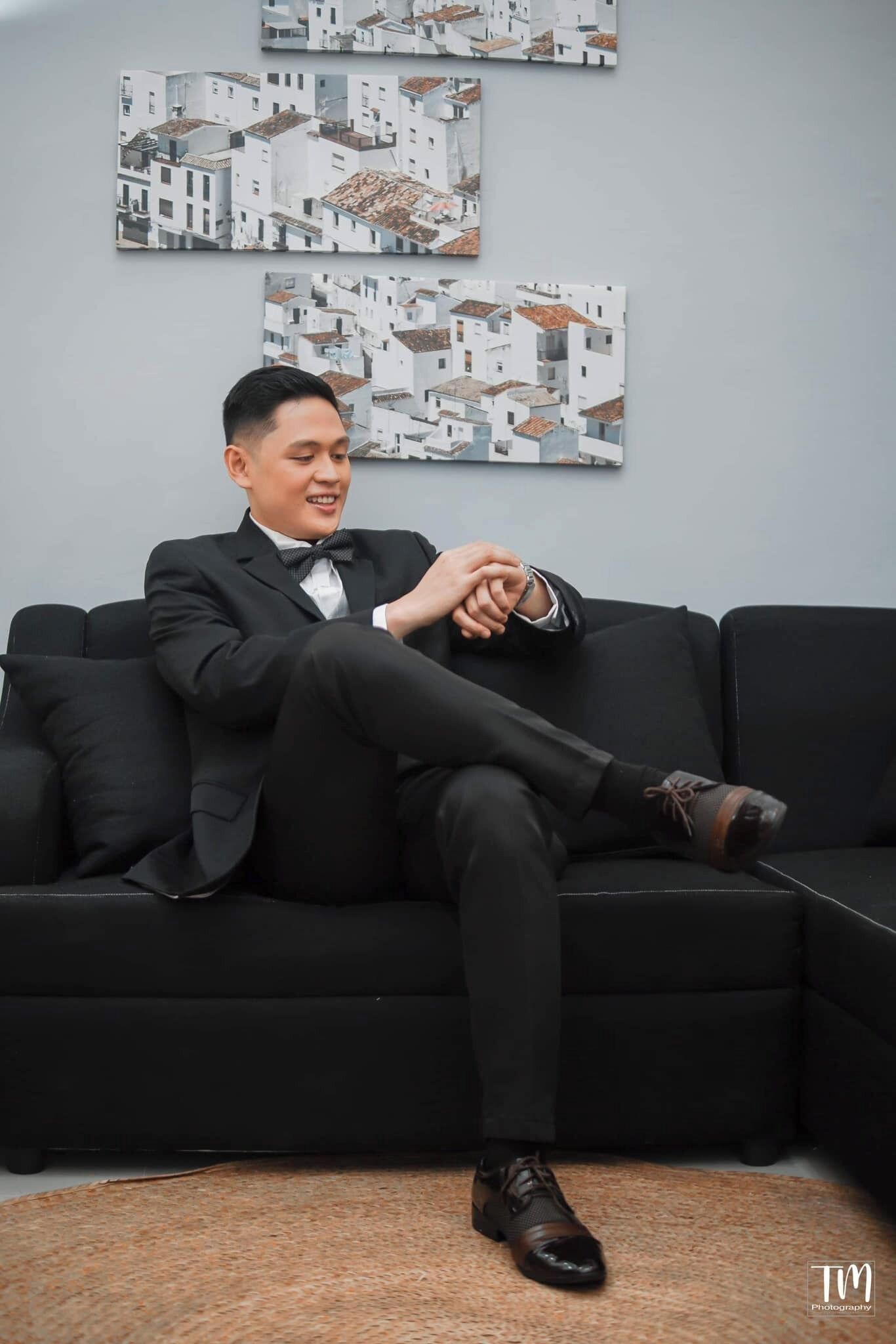 A person seated on a black sofa in a room with a light blue wall and two framed pictures depicting white buildings.