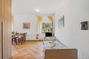 A room with a bed, a desk with chairs, a window with curtains, and a painting on the wall.