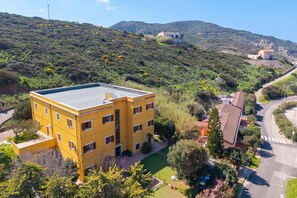 A yellow multi-story building with a flat roof, surrounded by greenery and hills in the background. - Residence Argentiera Studio 15 for four, Miniera dell’Argentiera, Italy (Miniera dell’Argentiera)