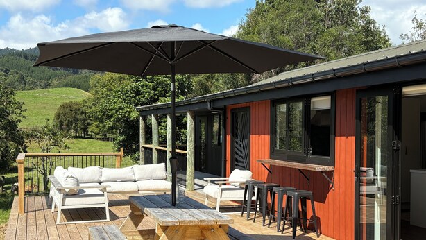 Une terrasse en bois avec des meubles d’extérieur, un grand parasol et une porte vitrée donnant sur un espace abrité.