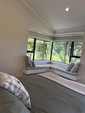 A bedroom with a large window, a built-in bench with pillows, and a bed. - Secluded, Relaxing 4-bedroom farmhouse in peaceful Ōrere (Ōrere Point)