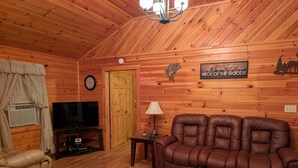 Ein gemütliches Blockhaus-Interieur mit Holzbalkendecke, einer Ledersofa, einem Fernseher und einem Schild mit der Aufschrift 'Welcome to our NECK OF THE WOODS'.
