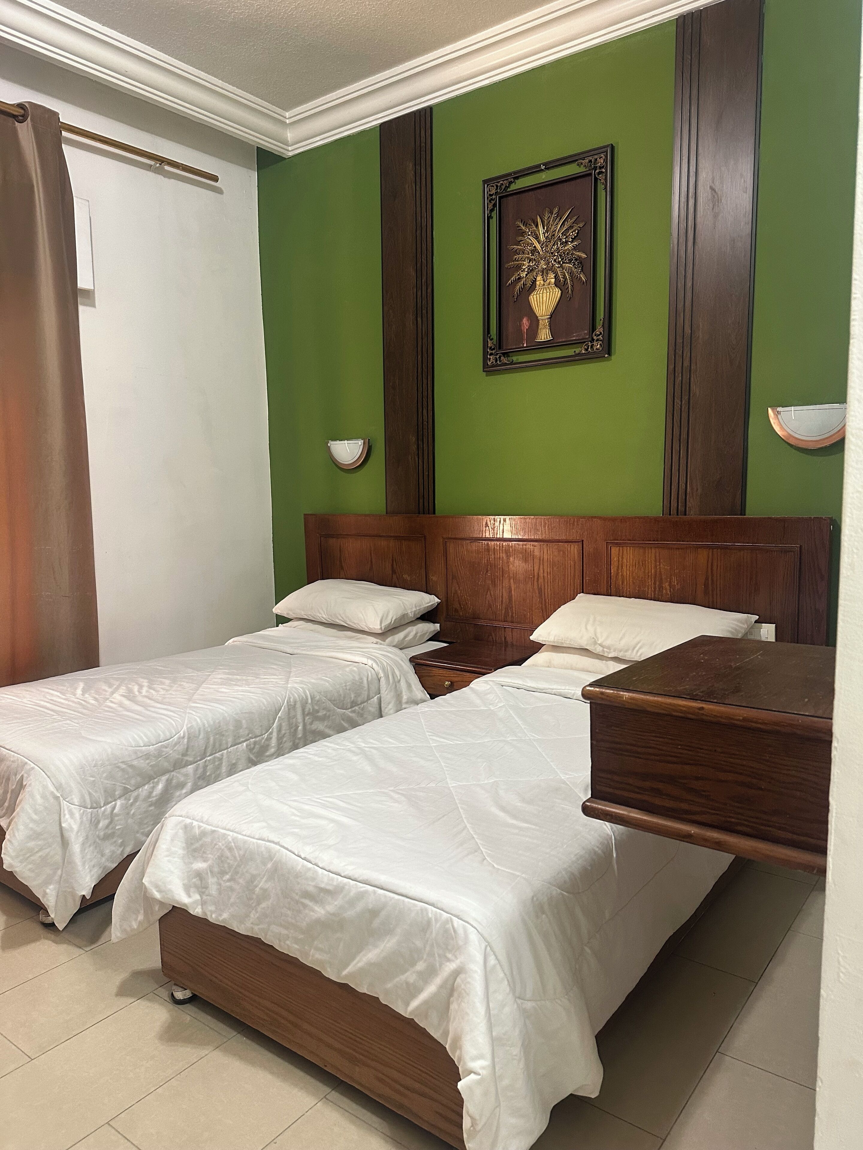 A hotel room with two single beds, a wooden headboard, a green accent wall, a framed picture, and a bedside table with a lamp.