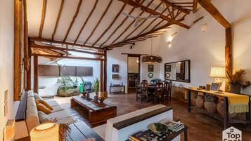 A spacious living area with a high ceiling, wooden beams, a sofa set, a dining table, and a kitchen in the background.