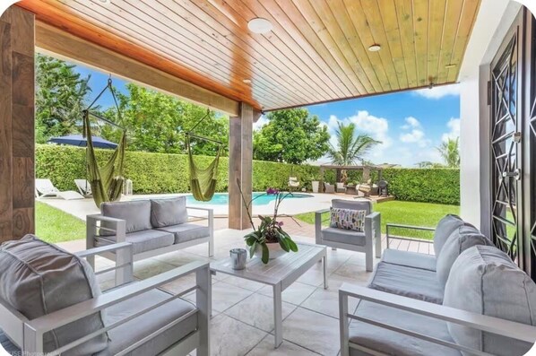 Un patio al aire libre con sofás grises y una mesa de centro blanca, techo de madera y vista a una piscina y follaje.