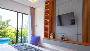 A modern hotel room with a large bed, a TV mounted on the wall, a small shelf with decorative items, and a view of a swimming pool. - Pondok Uma Jero (Tabanan)