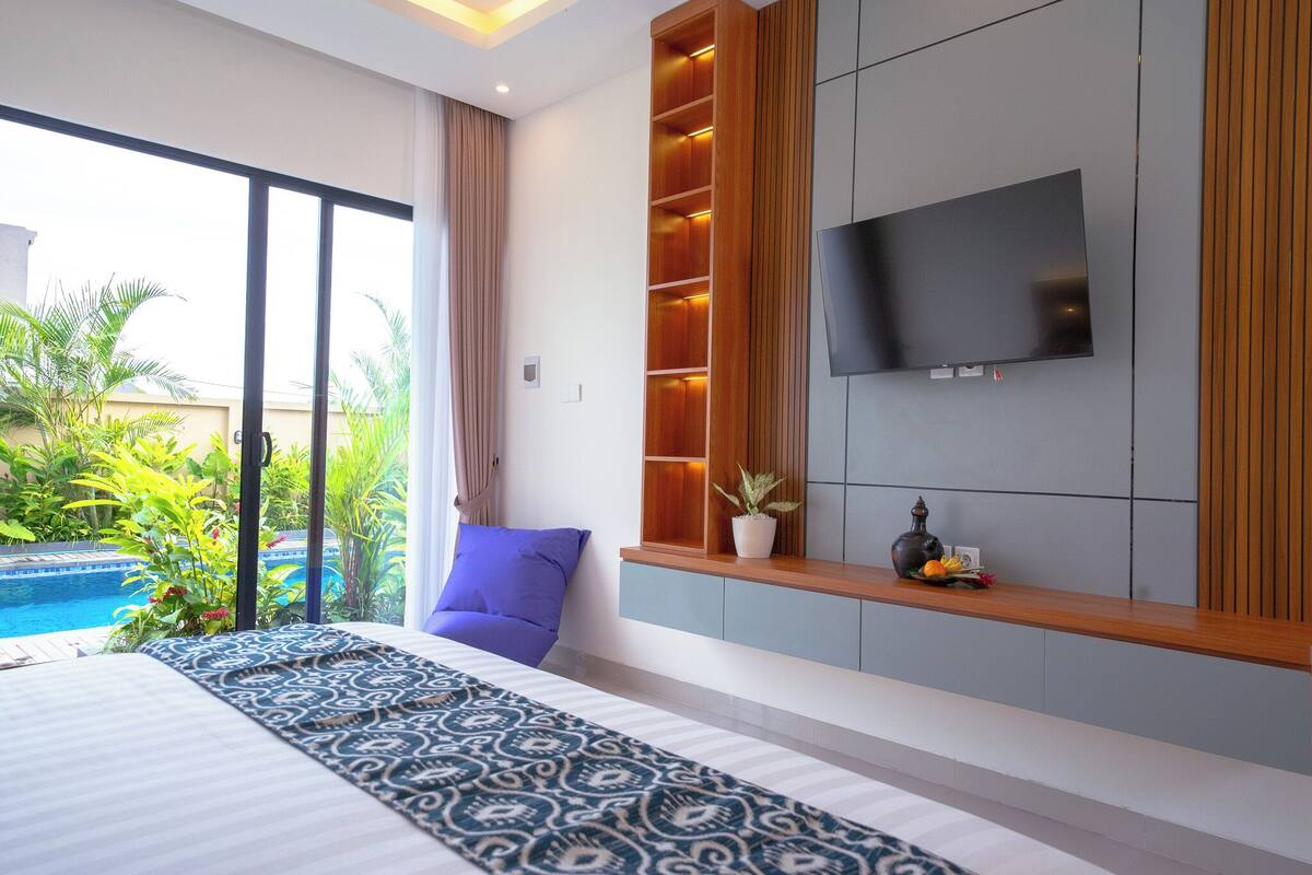 A modern hotel room with a large bed, a TV mounted on the wall, a small shelf with decorative items, and a view of a swimming pool.