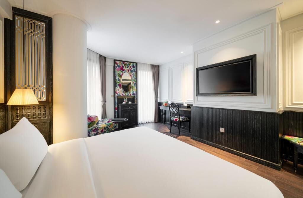 Ein Hotelzimmer mit einem großen Bett, einem an der Wand montierten Fernseher, einem Schreibtisch mit Stuhl und einem dekorativen Spiegel.
