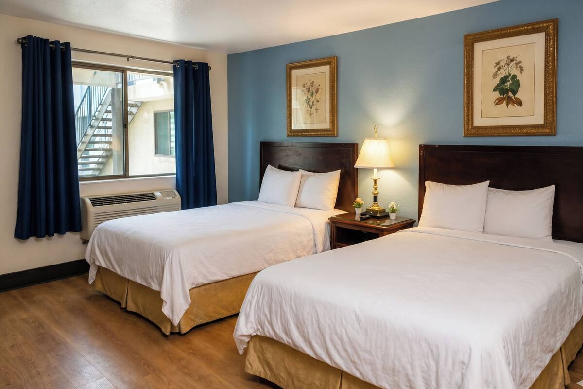 A hotel room with two beds, a window with curtains, a wall-mounted air conditioner, a nightstand with a lamp, and framed botanical prints.