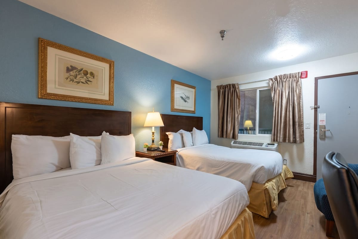 A hotel room with two beds, a window with curtains, a wall-mounted lamp, and framed pictures.