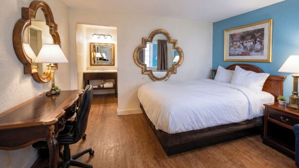 Quarto de hotel com escrivaninha de madeira, cama com roupa de cama branca, espelho e quadro emoldurado na parede.