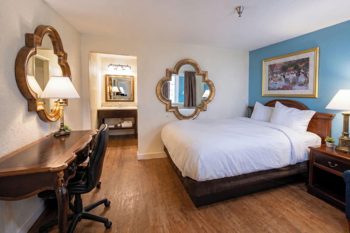 A hotel room with a wooden desk, a bed with white linens, a mirror, and a framed picture on the wall.