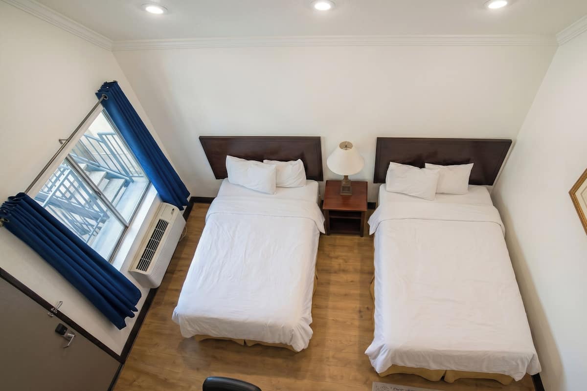 A hotel room with two beds, a window with blue curtains, a bedside table with a lamp, and an air conditioning unit.