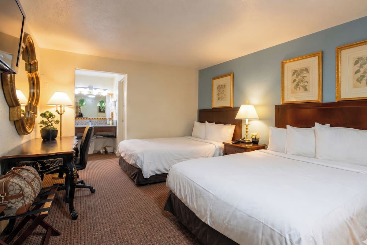 A hotel room with two beds, a desk with a chair, a lamp, and framed pictures on the wall.